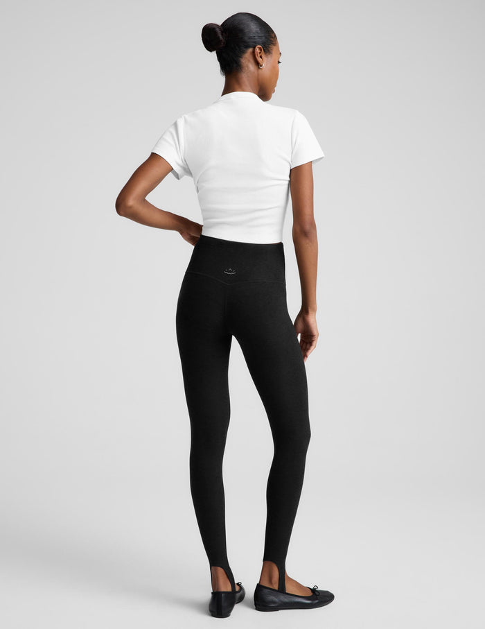 A woman modeling black leggings, a white top, and a black draped jacket, standing against a light gray studio background.
