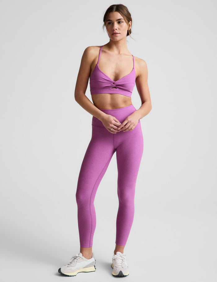 Woman wearing a purple sports bra and leggings on a light gray background.