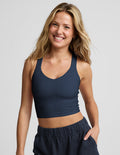 A smiling blonde woman wearing a dark navy blue crop tank and matching navy shorts, photographed against a solid light gray studio background.