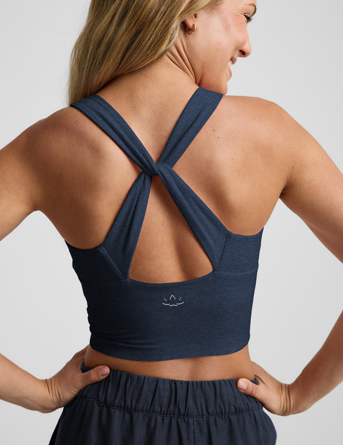 A woman shown from the back wearing a navy blue cropped tank with a crossover crisscross strap design and a small white logo on the lower center back, paired with matching dark bottoms against a light gray studio background.