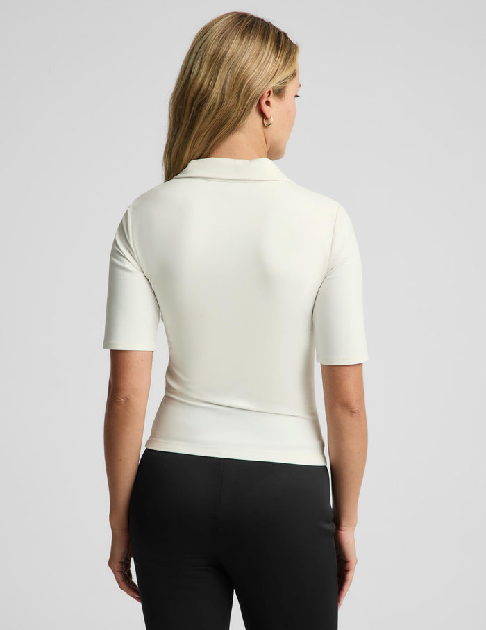 A woman shown from the back wearing a fitted short-sleeve polo-style top in off-white and black pants, photographed against a light gray studio background.