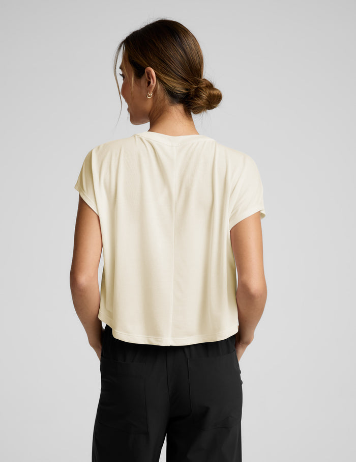Woman wearing a cream colored cropped tee and black pants against a plain background.