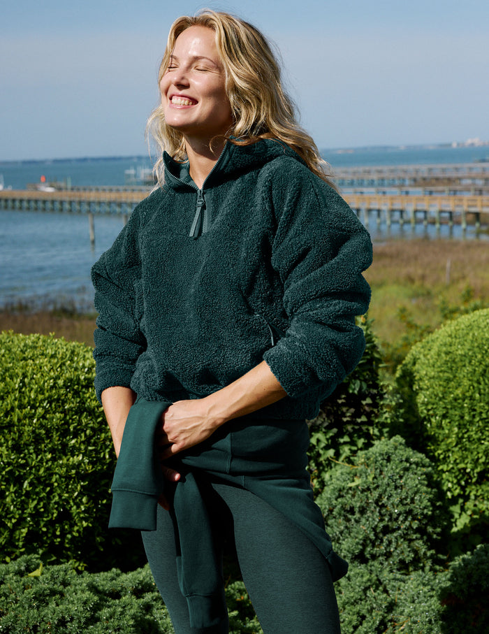 A woman outdoors wearing a dark forest green fleece pullover and matching green leggings, standing in front of green hedges with a pier and water in the background.
