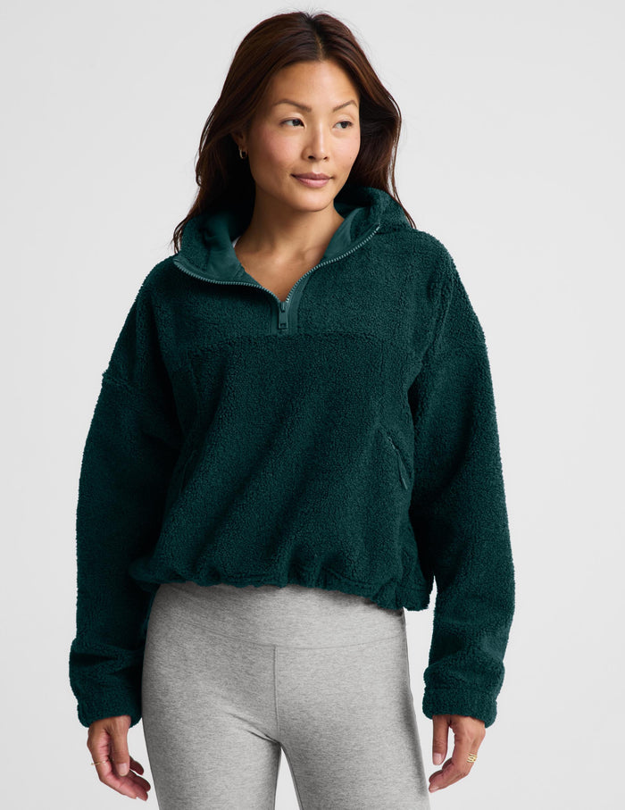 A woman wearing a dark green fleece half-zip pullover sweater and light gray leggings, standing against a light gray studio background.