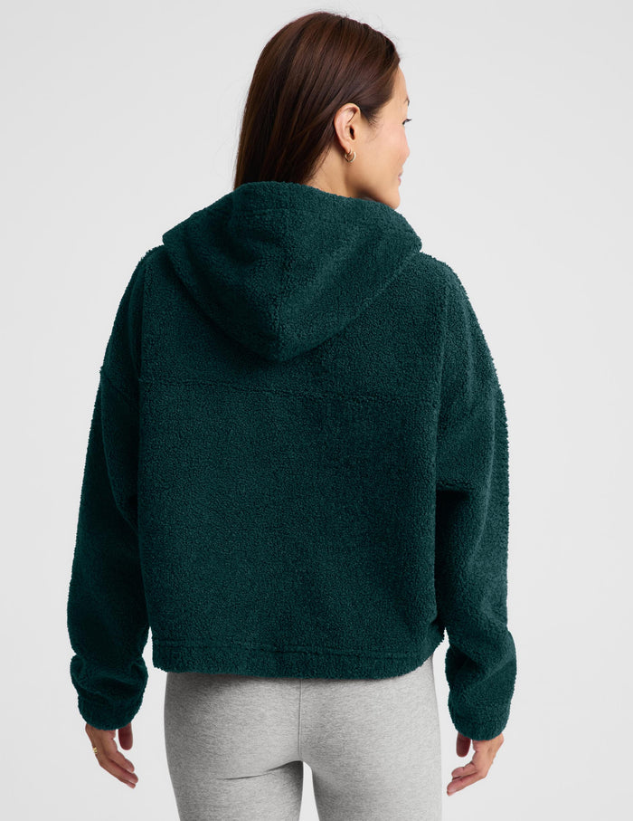 A woman wearing a dark green fleece half-zip pullover sweater and light gray leggings, standing against a light gray studio background.