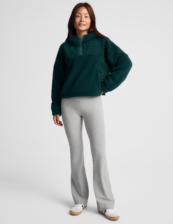 A woman wearing a dark green fleece half-zip pullover sweater and light gray leggings, standing against a light gray studio background.