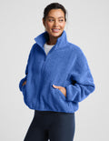 A woman wearing a textured fleece zip-up jacket in a medium blue color, paired with dark leggings, against a light gray studio background.