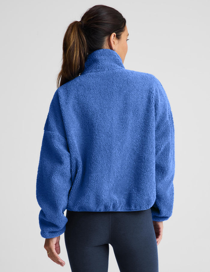 A woman wearing a textured fleece zip-up jacket in a medium blue color, paired with dark leggings, against a light gray studio background.