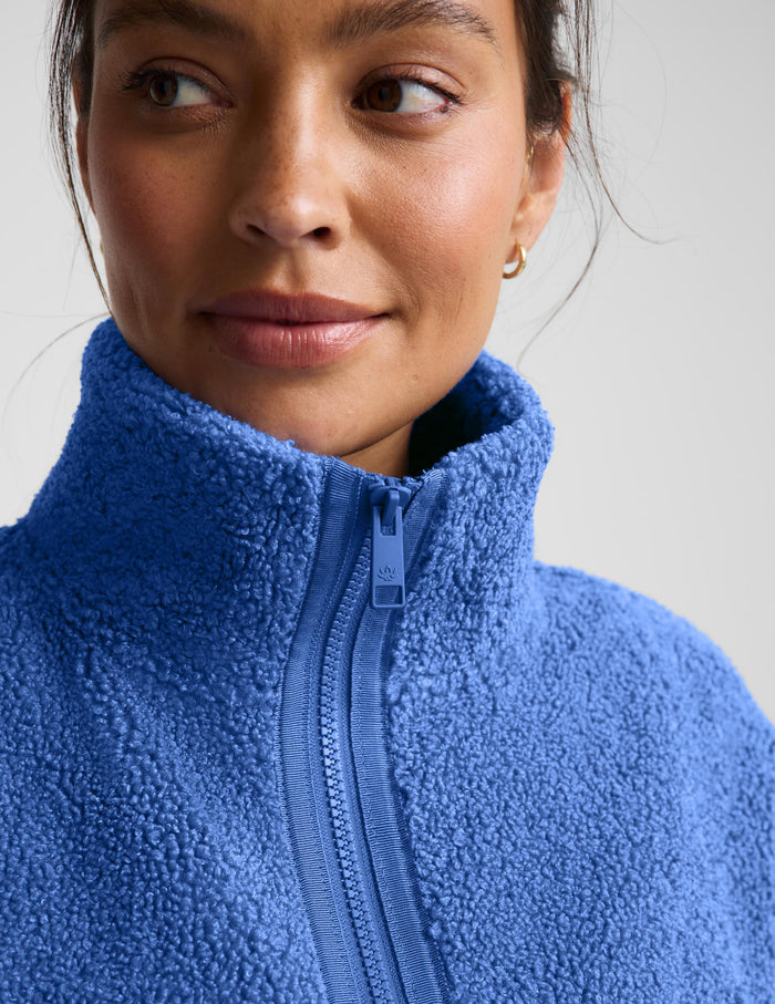 A woman wearing a textured fleece zip-up jacket in a medium blue color, paired with dark leggings, against a light gray studio background.