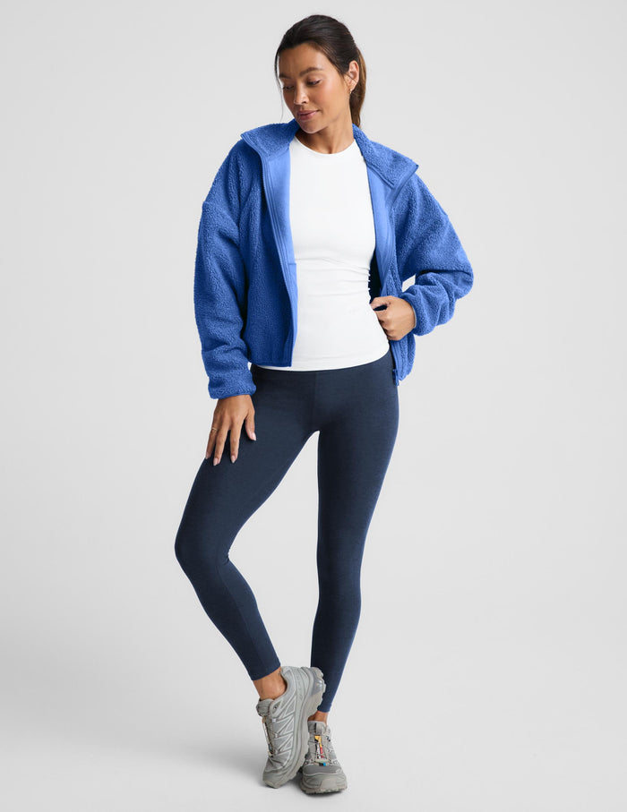 A woman wearing a textured fleece zip-up jacket in a medium blue color, paired with dark leggings, against a light gray studio background.